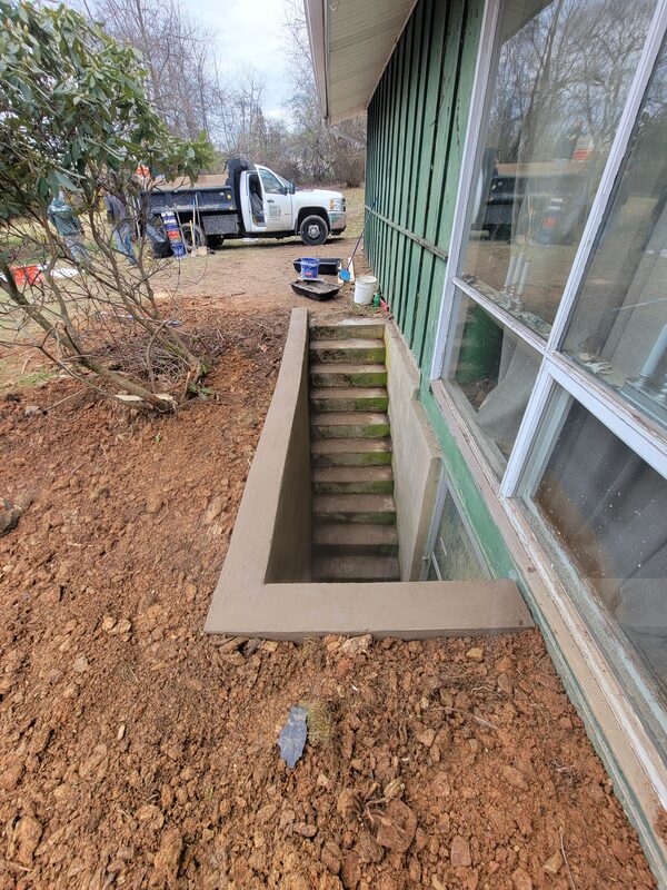 New concrete stairwell dug along the side of a house, leading down to a basement door, with fresh dirt around it and a work truck in the yard