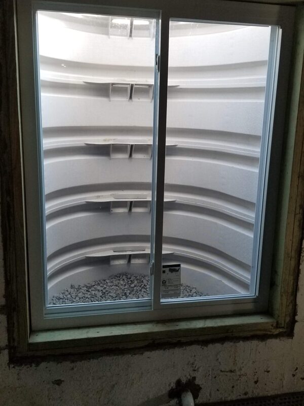Egress window seen from inside an unfinished basement, with a tiered window well outside and a pipe on the wall below