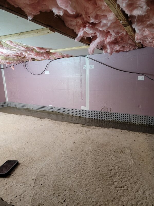 Fiberglass insulation sagging out of the floor joists above a damp floor, with water standing along the base of the wall