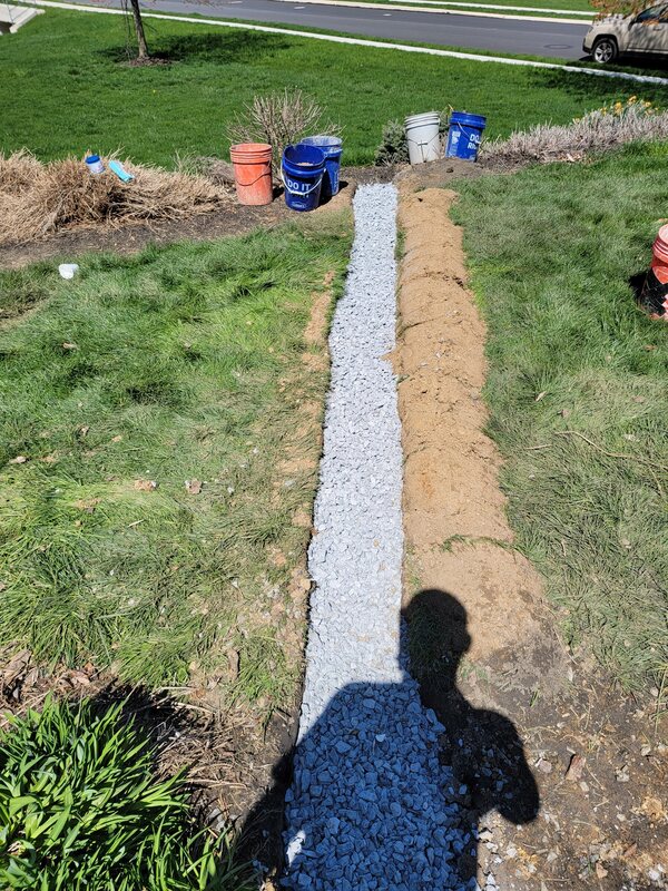 Shallow yard drain trench filled with clean gravel running across a lawn, with the excavated soil piled alongside