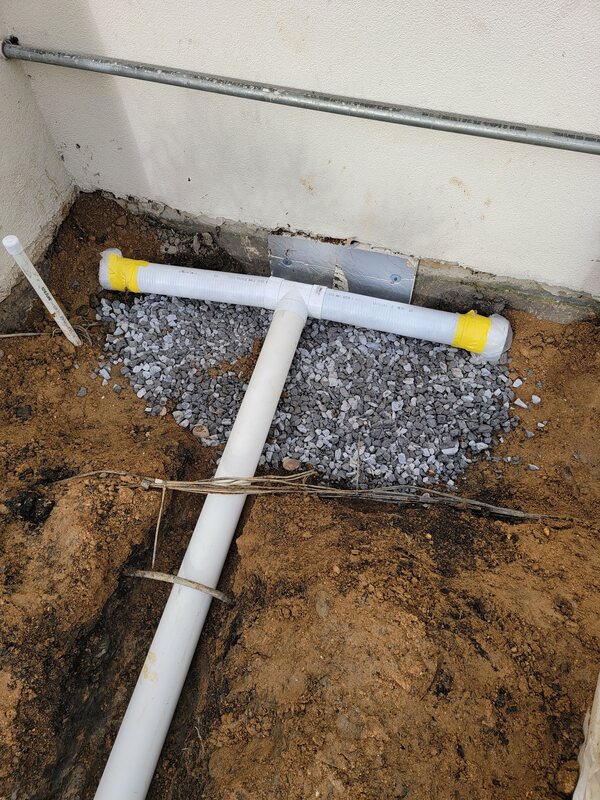 White PVC tee fitting set in a bed of clean stone at the base of a house wall, connected to a solid drain pipe running away from the foundation