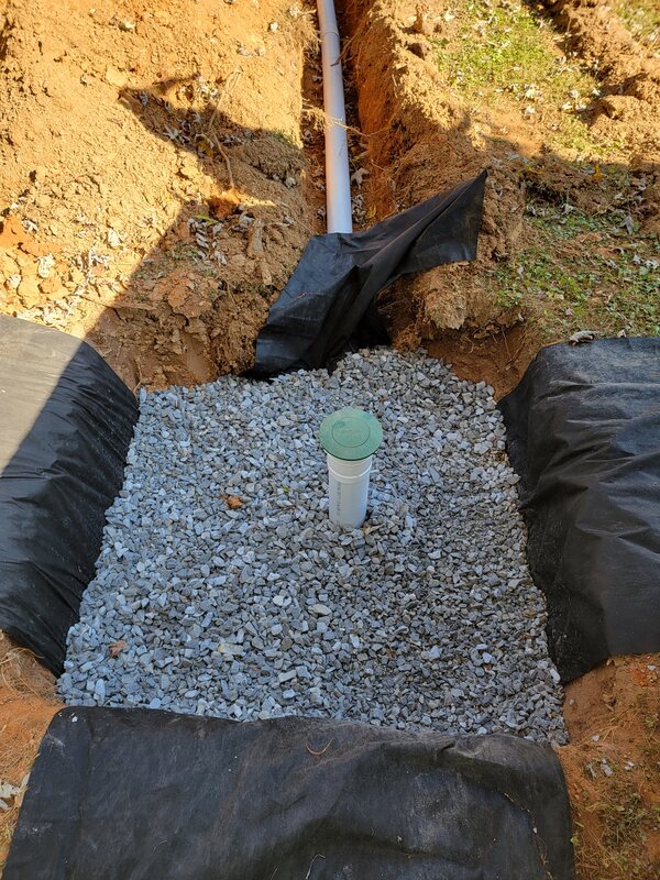 Dry well under construction: a square pit lined with black filter fabric, filled with clean gravel, with a green-capped inspection riser and an inlet pipe entering from the trench